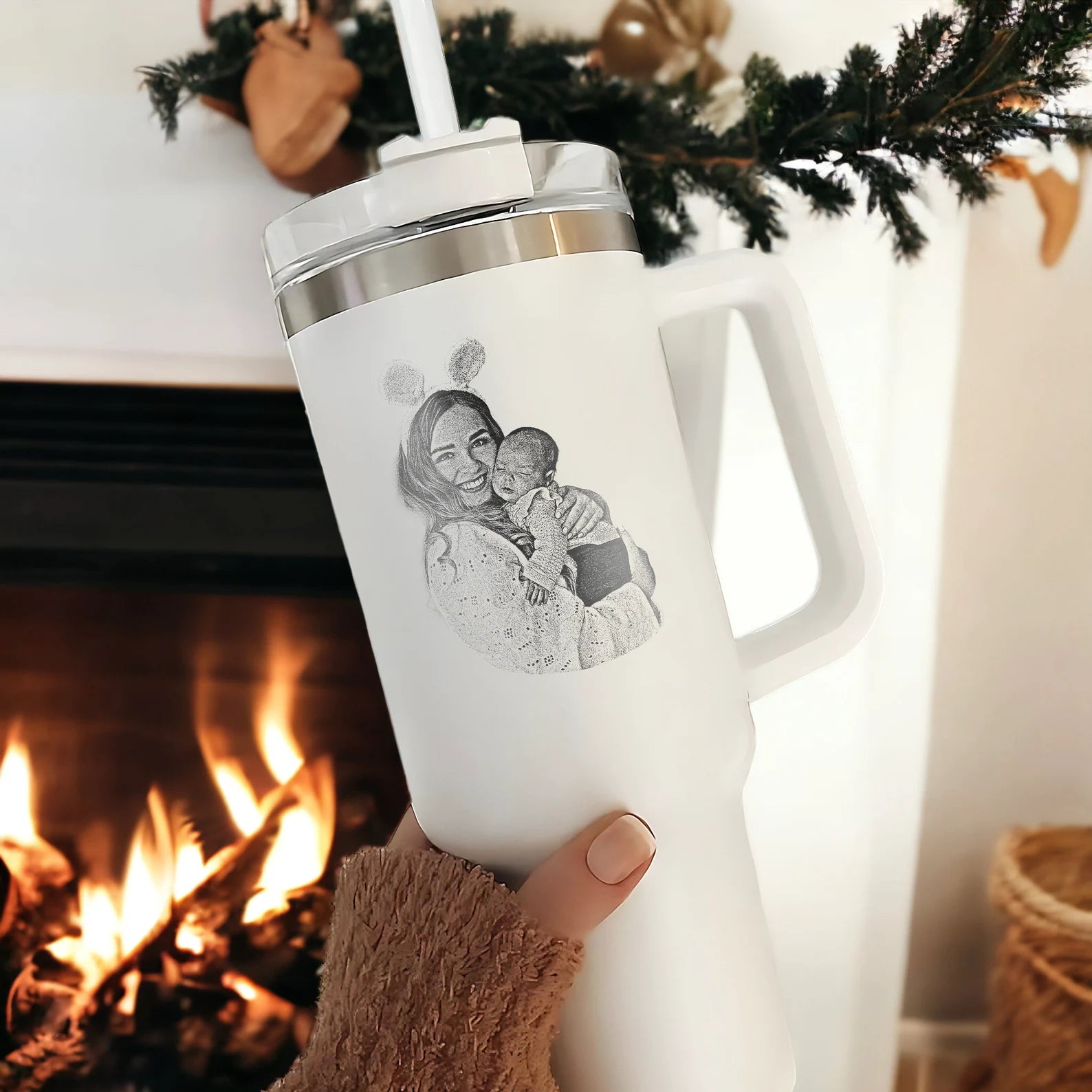 a large white tumbler similar to a Stanley brand cup that features an etched a custom photo of a woman and her baby; held by a feminine hand & set against a tidy background with a warm fireplace and holiday wreath