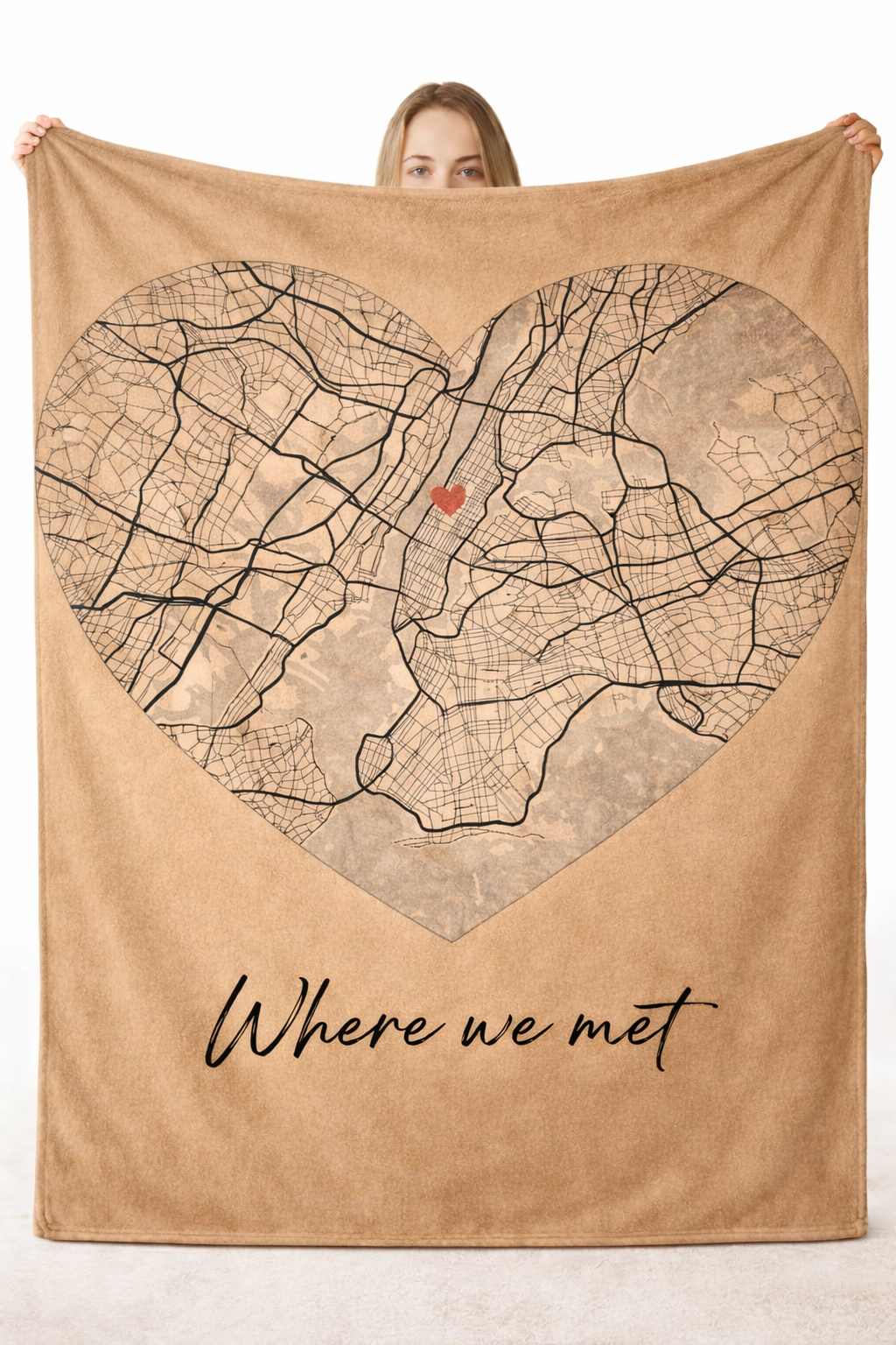 a woman holding up a large brown blanket featuring a map set in a heart-shaped frame and the text "Where we met"