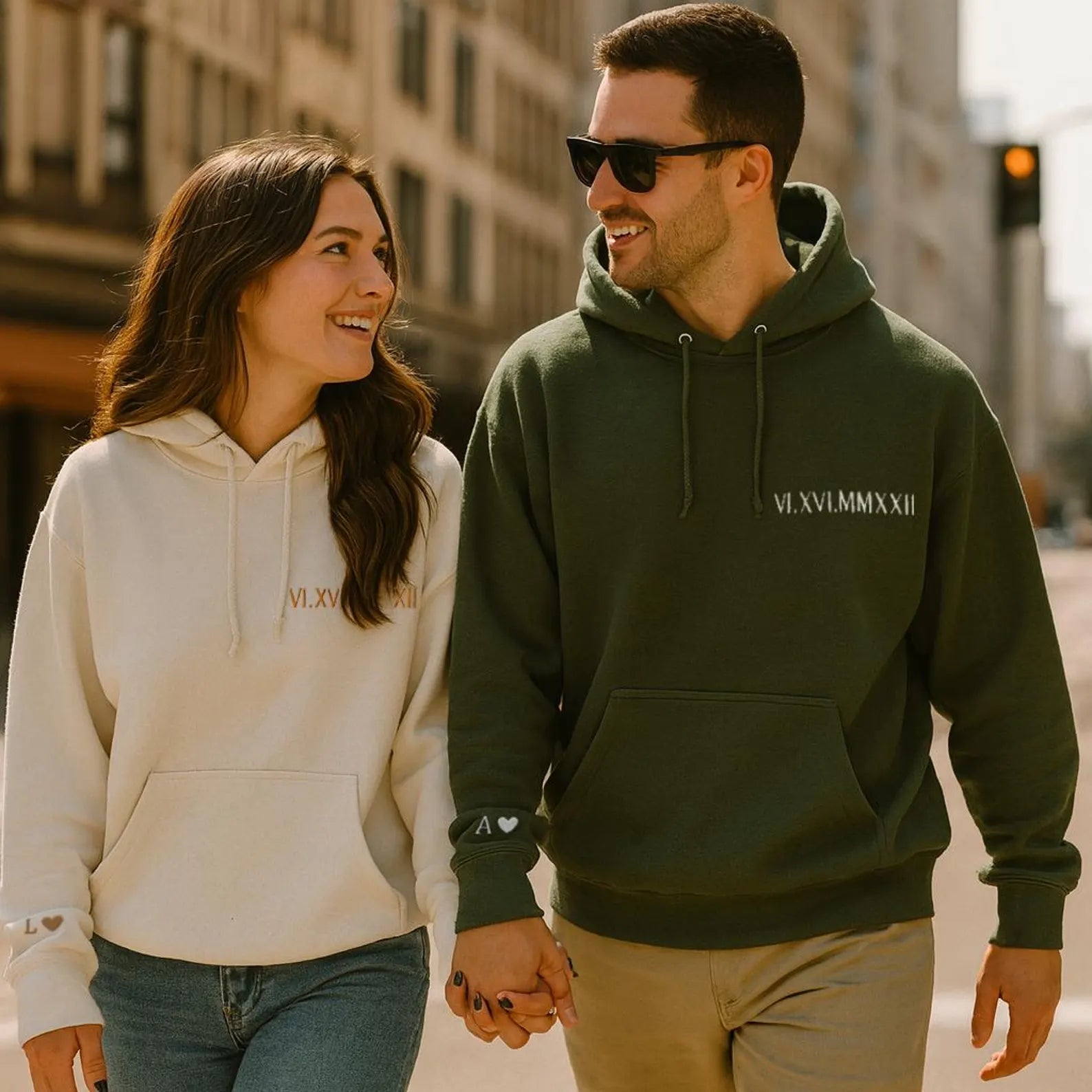 a smiling couple walking hand-in-hand wearing a cream-colored hoodie and a green hoodie, both featuring the same set of personalized roman numeral embroidery