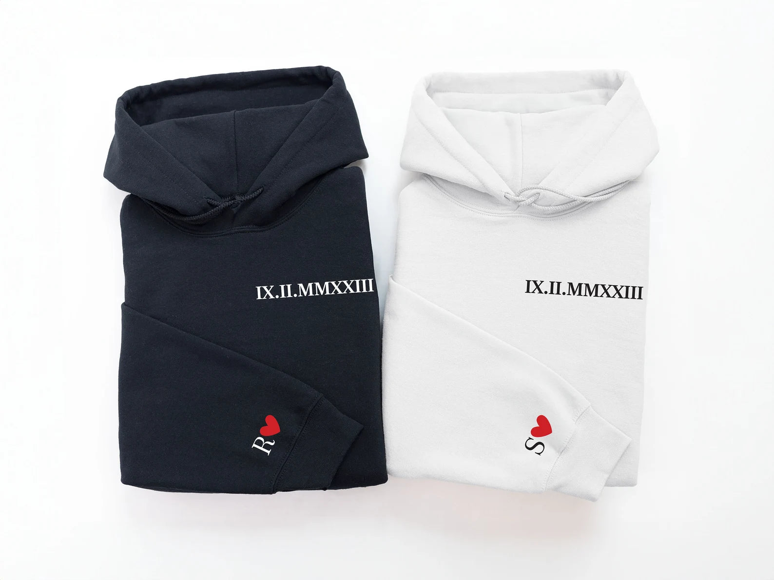 a black hoodie and a white hoodie folded neatly side-by-side, each featuring the same roman numeral on the left chest and a different initial on the sleeve cuff