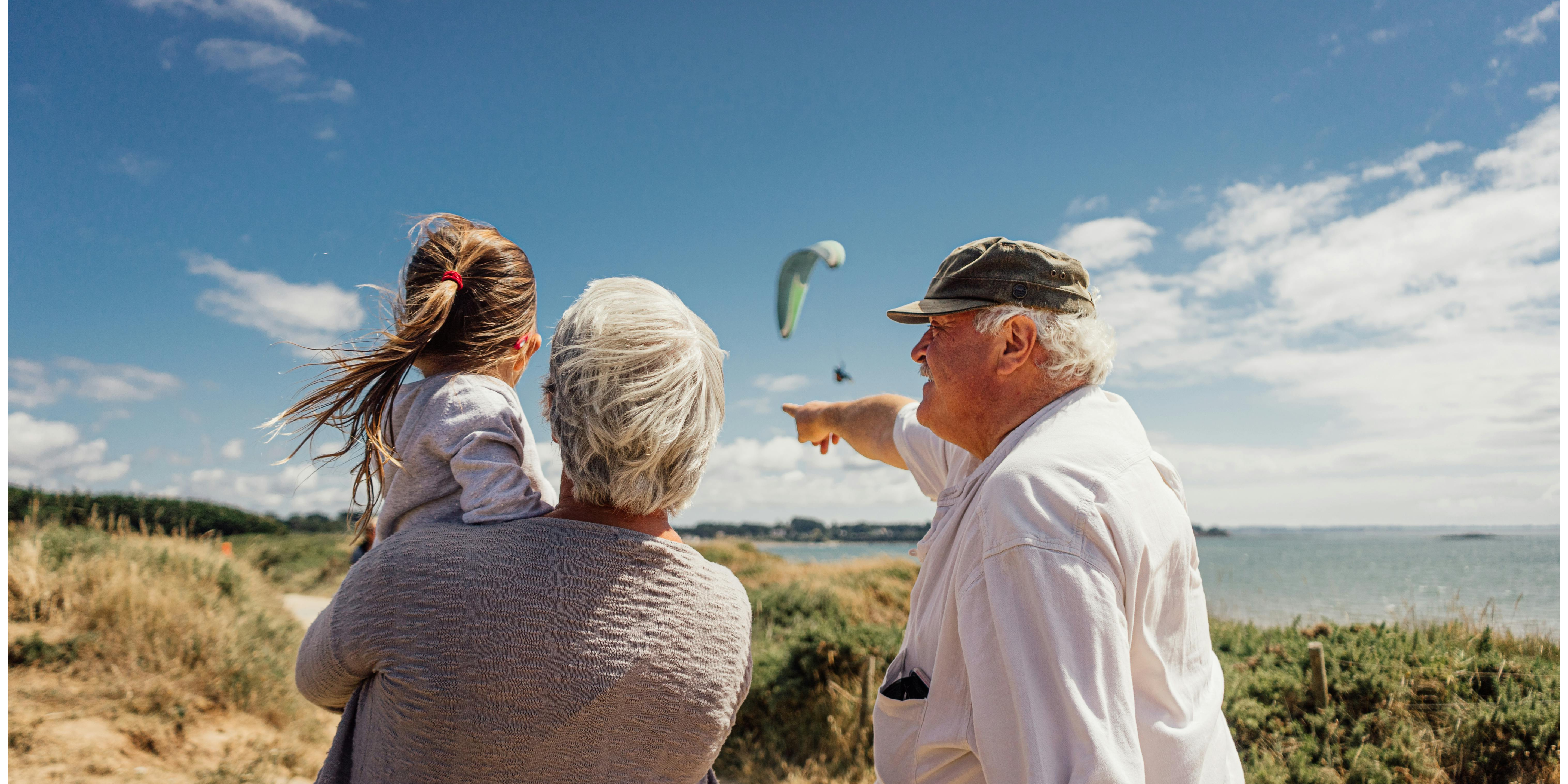 an elderly couple out on the beach with their granddaughter. the man is smiling at the child and pointing out a paraglider in the distance.