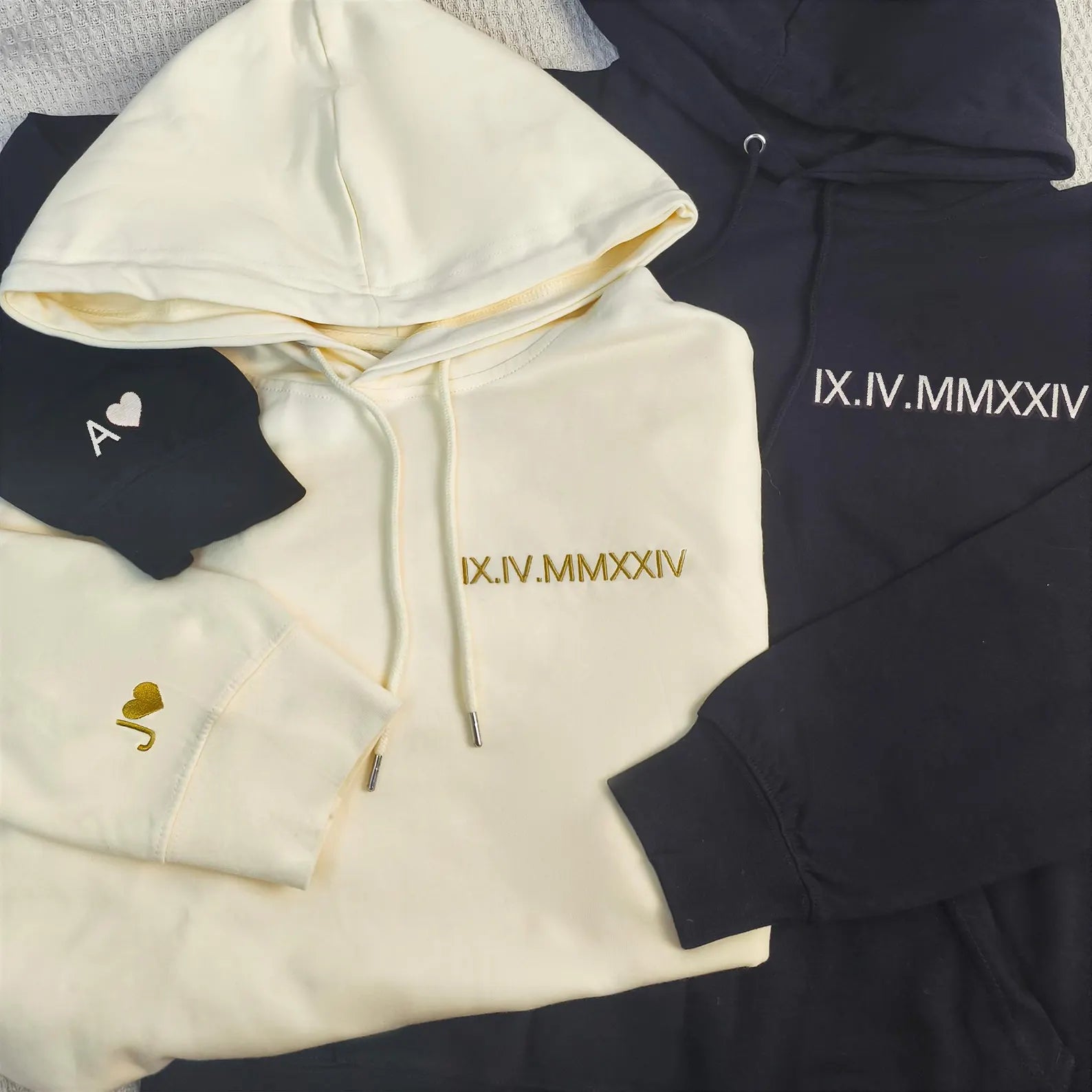 a cream-colored hoodie and a black hoodie folded neatly together and each featuring the same set of roman numeral embroidery