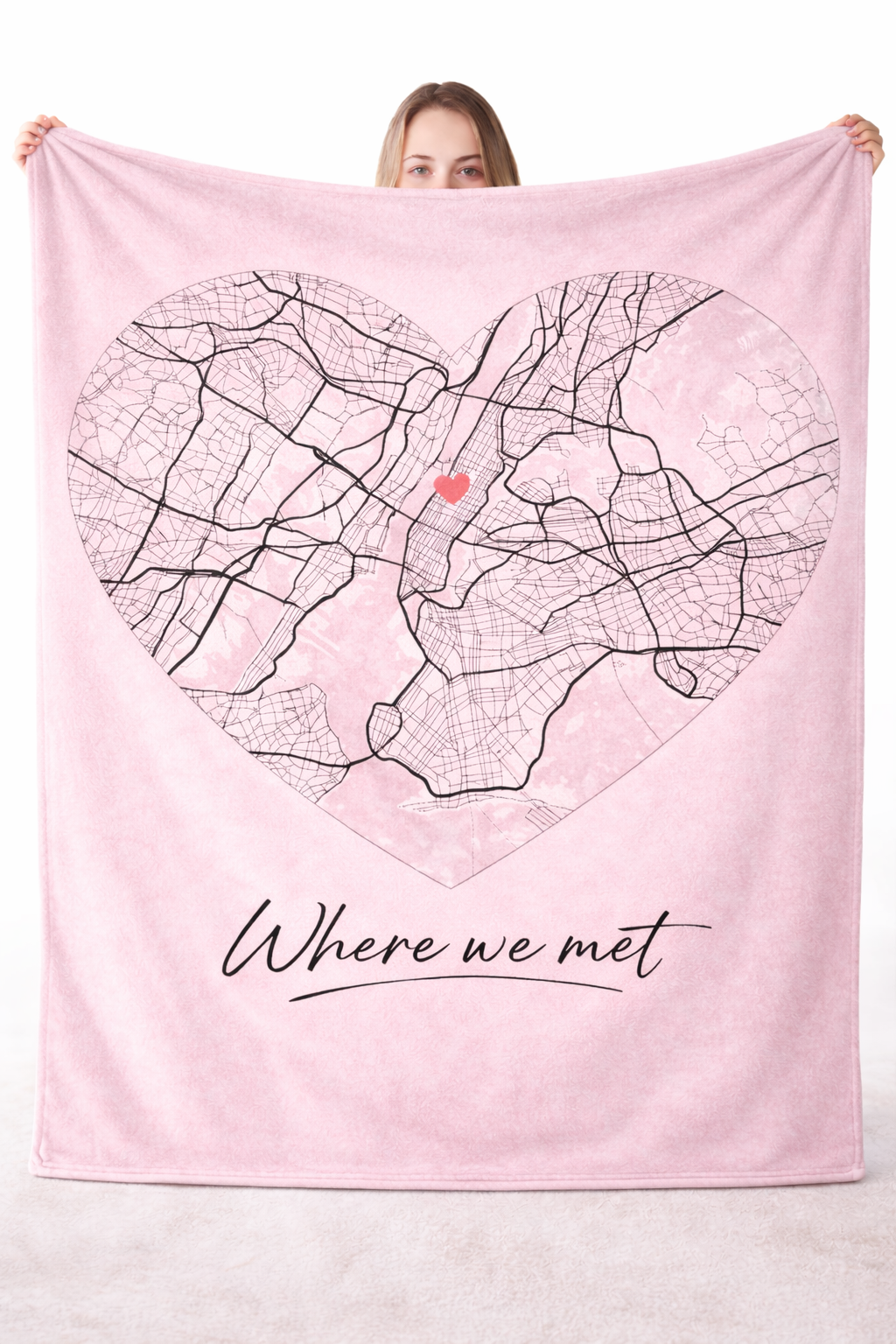 a woman holding up a large pink blanket featuring a map set in a heart-shaped frame and the text "Where we met"