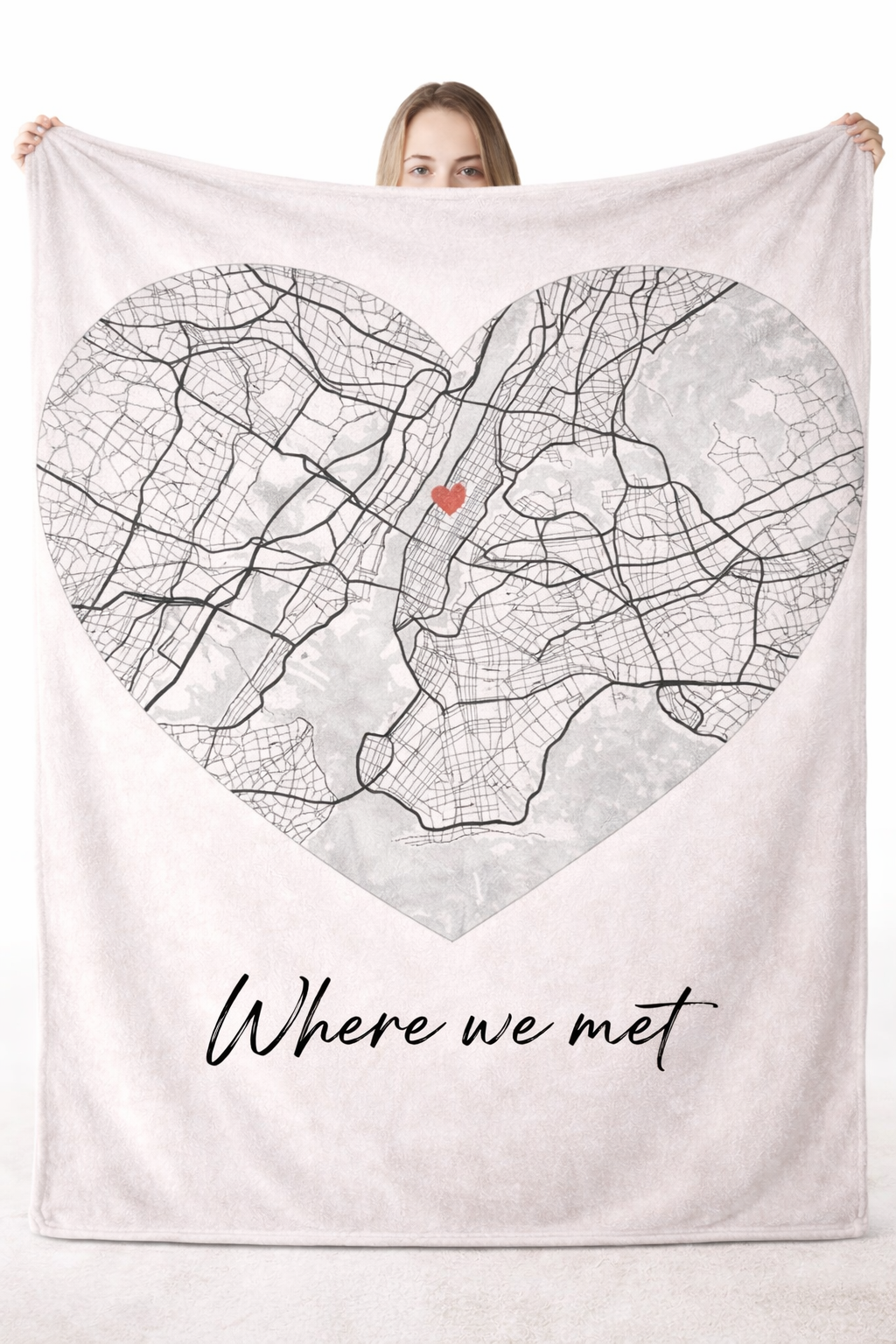 a woman holding up a large white blanket featuring a map set in a heart-shaped frame and the text "Where we met"