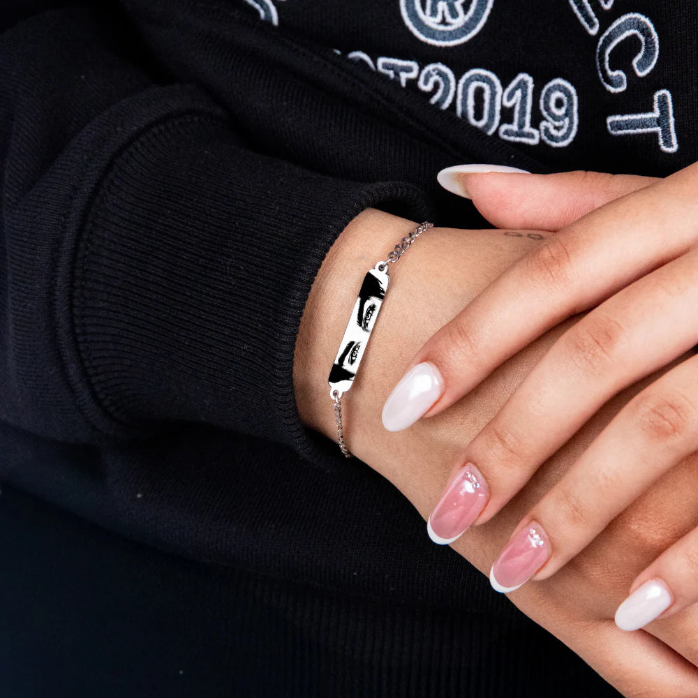 a silver bracelet featuring a printed greyscale photo of a man's eyes is worn by a woman with painted white and pink nails in this closeup image