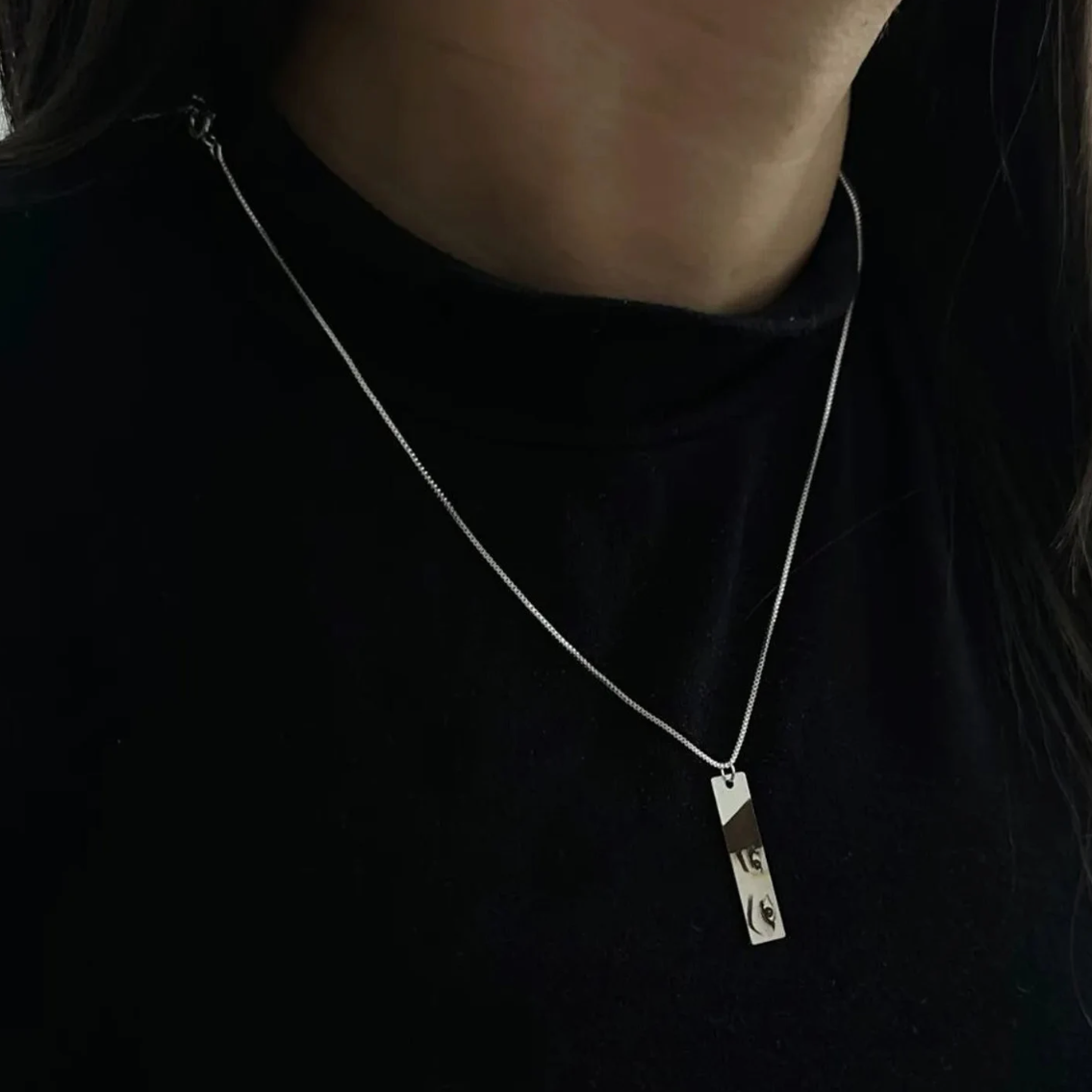 a closeup of a woman in a black t-shirt wearing a rectangular silver necklace pendant with a printed photo of another young lady's eyes in black and white