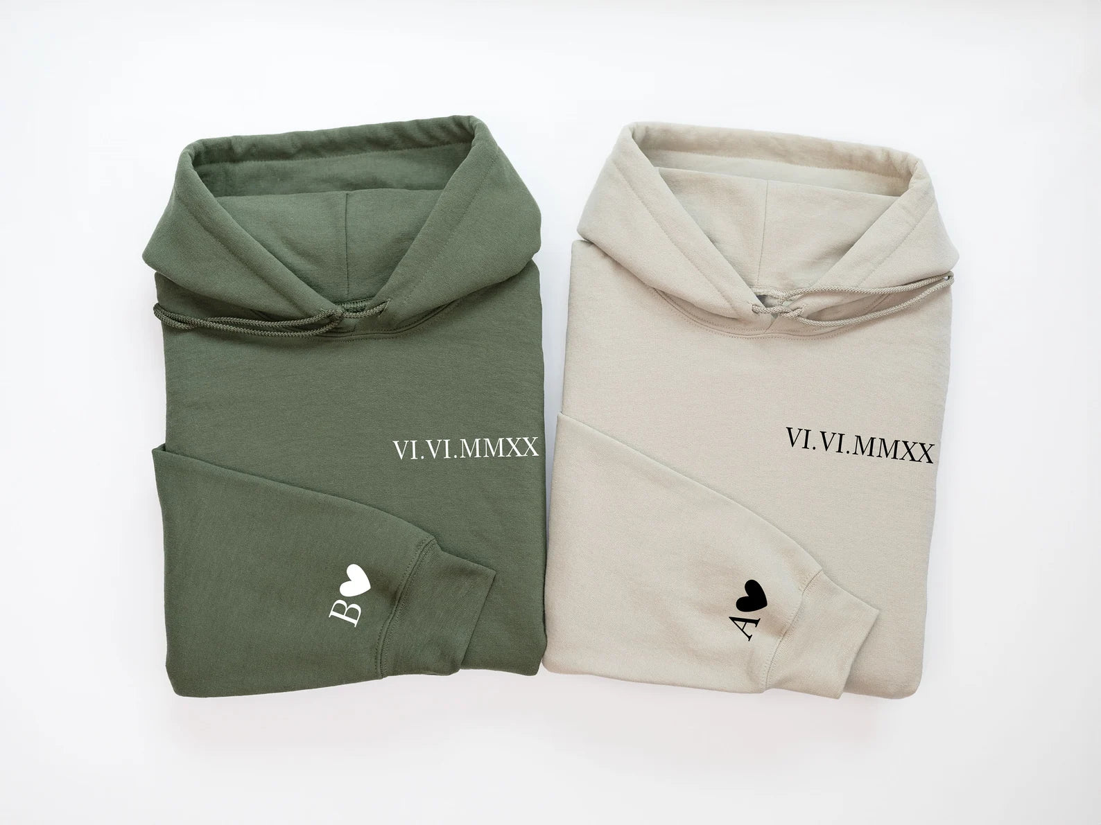 a cream-colored hoodie and a pale green hoodie folded neatly together and each featuring the same set of roman numeral embroidery on the left chest with different initials on the sleeves