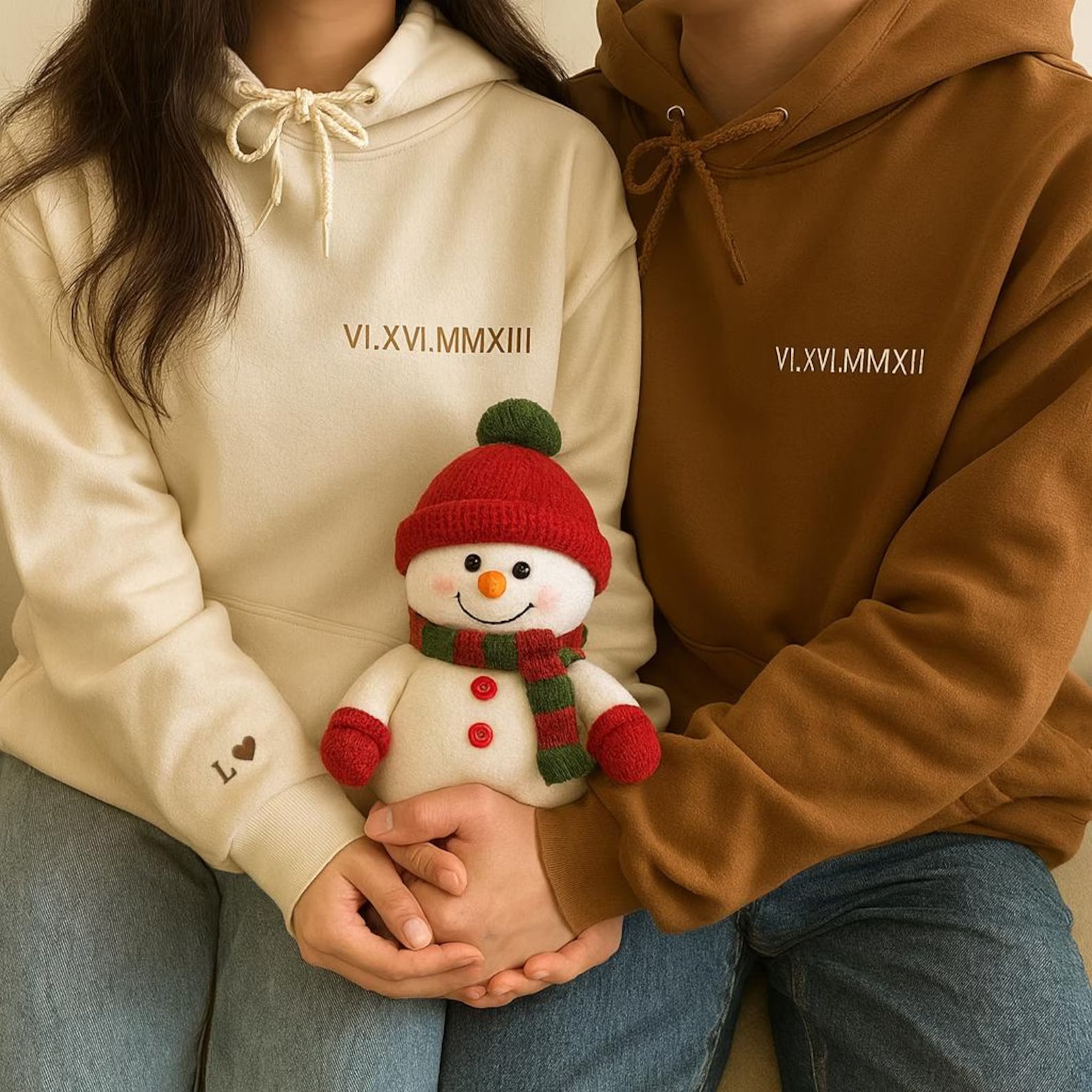 a woman and man wearing a cream-colored hoodie and a brown hoodie, each featuring the same set of roman numeral embroidery; holding a smiling snowman plushie
